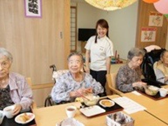 和やかな食事の様子 日常風景 明るい笑顔のスタッフが立つ食堂で、利用者同士が和気あいあいと食事を楽しんでいます。心地よいコミュニケーションが日々の喜びを支えています。