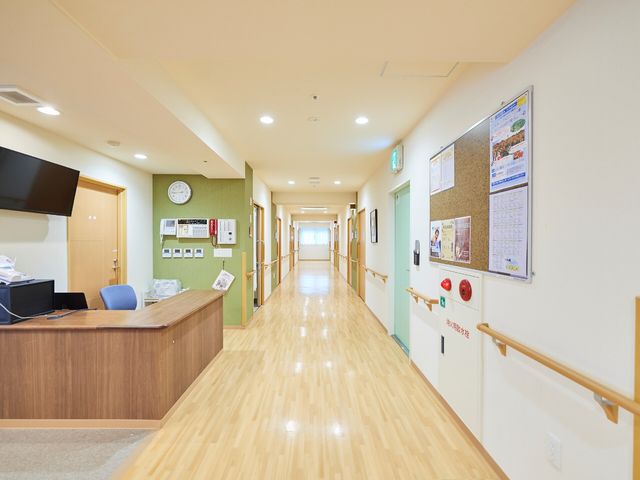 明るく清潔な廊下 ヘルパーステーション 手すりが設置された温かみのある広々とした廊下で、安心してお過ごしいただけます。フロントの横には、情報掲示板があります。