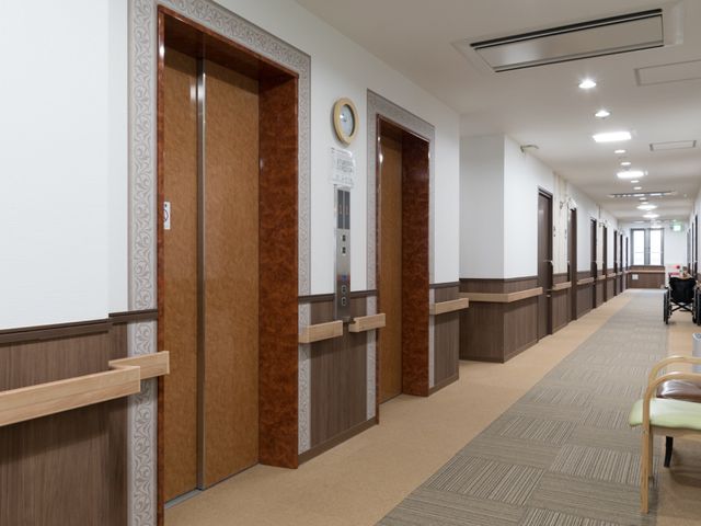 廊下とエレベーターの風景 エレベーター 清潔で明るい廊下は、手すりが整っており安全に移動できる設計です。背後には車椅子の方も見受けられます。