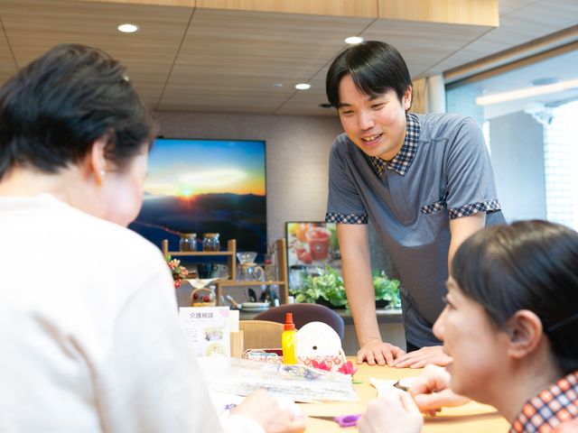 相談中の職員と利用者がいるラウンジ ラウンジ   自然光が入る明るい空間で、職員が利用者の方と向き合って相談しています。落ち着いた雰囲気で、会話しやすい場に見えます。