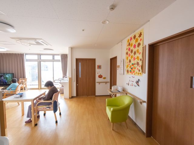 快適な居室の様子 リビング ゆったりとした明るいリビングで、利用者がテレビを観たり、テーブルでくつろいでいます。壁には季節感を感じさせる装飾が施され、居心地の良い空間となっています。