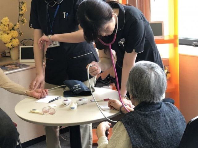 検診する職員と利用者 スタッフイメージ 和やかな雰囲気の中で、スタッフが利用者の健康チェックをしています。明るい空間で安心感を提供しています。