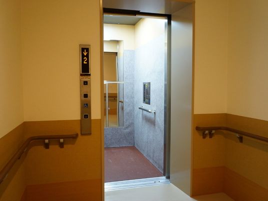 エレベーターが開いている様子。手前の壁は3色のグラデーションになっていて、茶色の手すりが付いている。エレベーターの中には鏡が見える。