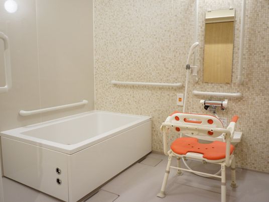 細かなモザイクタイルの壁の浴室。手すりがたくさんついている。シャワー、バスタブ、オレンジの肘かけ付き入浴イスが設置されている。