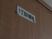サムネイル 白いプレートが居室のドアに設置されている。下部にはプラスチックのフックが2つついており、黒い文字で1丁目8番地と書いてある。