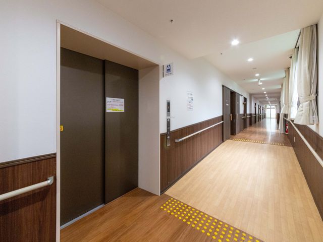 明るい廊下とエレベーター前 廊下   手すりが続く明るい廊下で、床の色分けや点字ブロックが見えます。エレベーター前も広く、移動しやすい印象です。