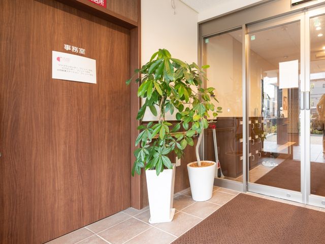 エントランスの植物と案内板 玄関 エントランスは清潔感のある設計で、温もりを感じる案内板が訪問者を歓迎しています。植物がある明るい受付は、来訪者に心地よい印象を与えます。