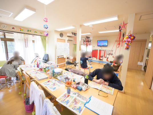 施設の写真 ダイニングテーブルが一列に並べられたフロアがあり、奥にはトレーニング機器が置かれている。入居者様がスタッフと共に身体を動かしている。