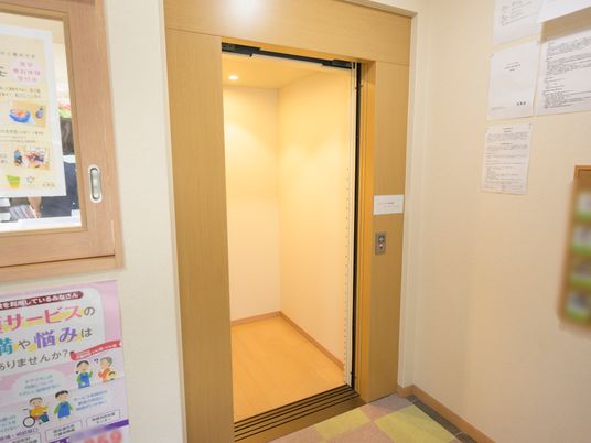 施設の写真 フロアの一角にエレベーターが設置されている。扉や床が木目調になっていて、温かみのある印象を与えている。