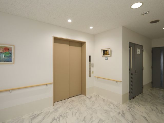 廊下とエレベーターの画 エレベーター 広々とした廊下は明るく清潔感があり、壁に掛けられた絵画が訪れる人々を温かく迎えています。エレベーター前の空間もゆったりとしており、快適にお過ごしいただける環境が整っています。