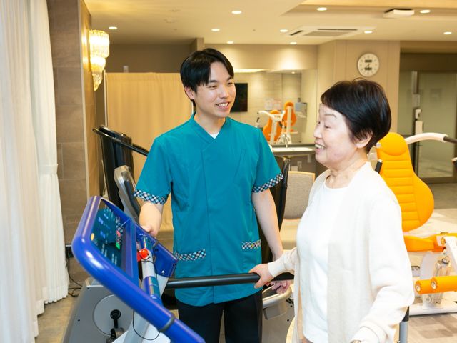 機能訓練室で談笑する様子 機能訓練室   明るい機能訓練室で、職員が利用者の方と笑顔で会話しています。運動器具が整い、働く現場の雰囲気が伝わります。