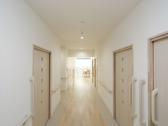明るい廊下と部屋の扉 廊下 手すりが備えられた広々とした廊下は、ご高齢の方々が安心して歩行できる配慮がされています。明るい窓からは心地よい陽光が差し込み、温かい雰囲気を演出しています。
