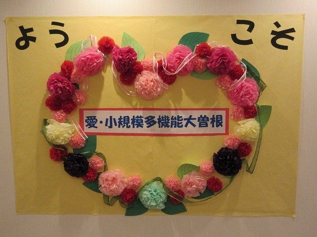掲示物 掲示物   黄色い台紙に、花飾りで作った輪と「ようこそ」の文字が飾られています。施設のあたたかな雰囲気が伝わる掲示です。