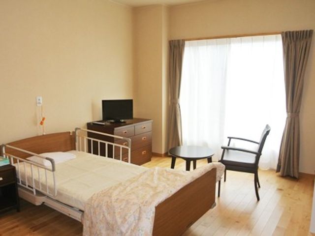 明るい居室のベッド 居室   大きな窓から光が入る、落ち着いた雰囲気の居室です。ベッドやテレビ台、丸テーブルがあり、ゆったり過ごしやすい空間です。