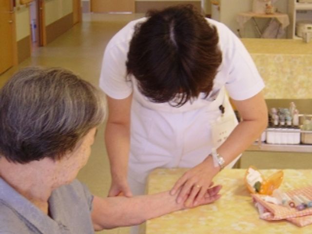 スタッフと利用者の対話 居室 ケアの様子が伝わる居室で、スタッフが利用者の手に優しく触れながら会話をしています。安心して過ごせる空間です。