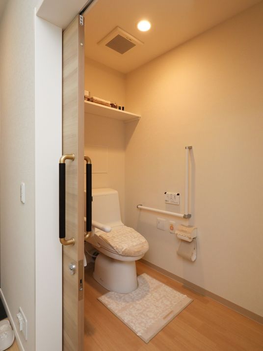 白い洋式トイレの蓋にカバーが掛けられ、床には揃いのマットが敷いてある。壁に手すりとリモコンとペーパーホルダーが設置されている。