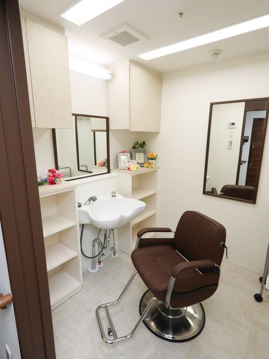 シャワー台の前に茶色い回転いすがある。椅子の正面と左側の壁に鏡が設置され、天井の蛍光灯が点灯している。