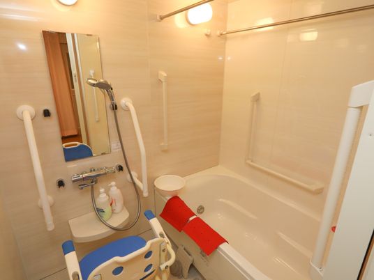 ユニットバスタイプの浴室。浴槽に２枚の赤いマットが掛けられている。洗い場に青いシャワー椅子が置いてある。