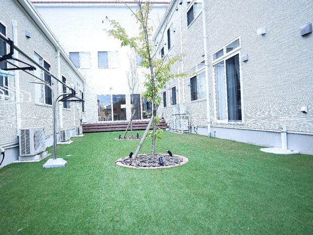 白壁の建物と緑の中庭 中庭 静かな緑の中庭は、日々の忙しさを忘れさせる癒しの場所です。屋外での憩いやレクリエーションにも最適です。