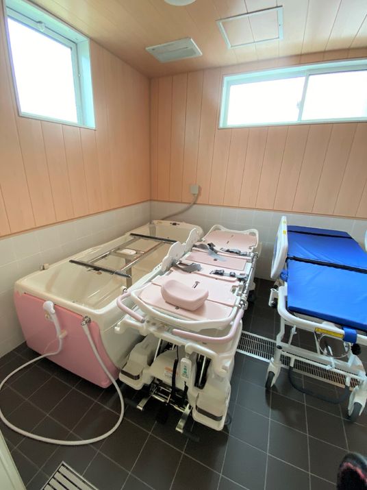白とピンクの寝台浴が設置された浴室。大きな窓があり、明るい雰囲気。ブルーのストレッチャーも機械の横に置かれている。