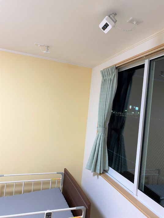 介護ベッドが置かれた居室には緑のカーテンが付いた窓が設置され、窓に近い天井部分にはセンサーカメラのようなものが付いている。