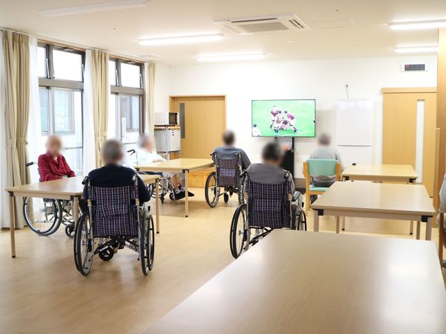 車椅子の利用者がいる食堂 共有スペース 広々とした食堂で、車椅子の方々がくつろぎながらテレビを楽しんでいます。共有の食事スペースは清潔感があり、くつろぎの時間を提供しています。