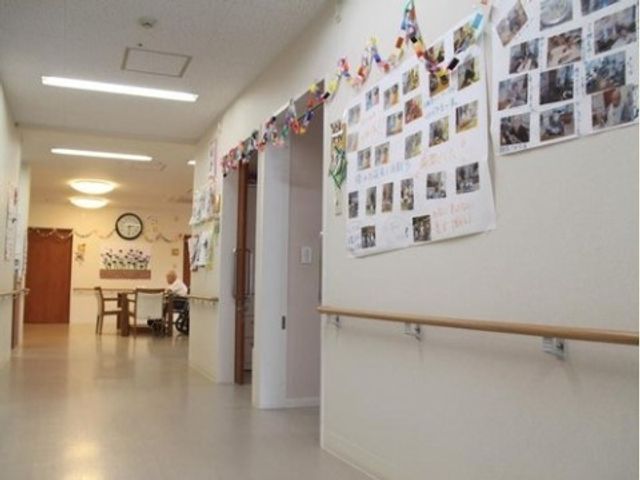 廊下に掲示物と座る人 廊下 手すり付きの廊下には様々な掲示物が飾られ、明るい印象を与えています。遠くの椅子には一人がくつろいでおり、生活の一コマを垣間見ることができます。
