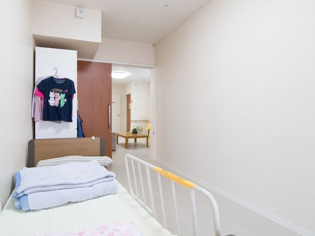 居室のベッドと扉 居室 清潔感のある居室のベッドには、整えられたリネンが置かれ、快適な休息が約束されます。扉の向こうには共有スペースが広がり、暖かいコミュニティの雰囲気を感じさせます。