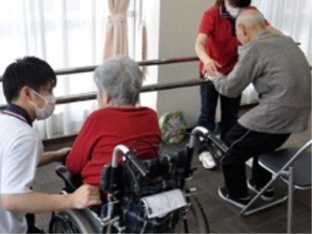 活動支援の場面 リハビリ 職員が高齢者の体を支えながら立ち上がりの練習を見守っています。経験豊かなスタッフと共に、利用者の自立をサポートする穏やかな環境です。