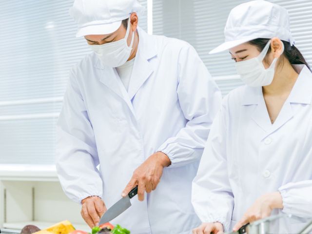 調理をするスタッフの様子 スタッフイメージ 調理服を着たスタッフが、清潔な環境で料理を準備しています。職場の衛生管理が行き届いています。