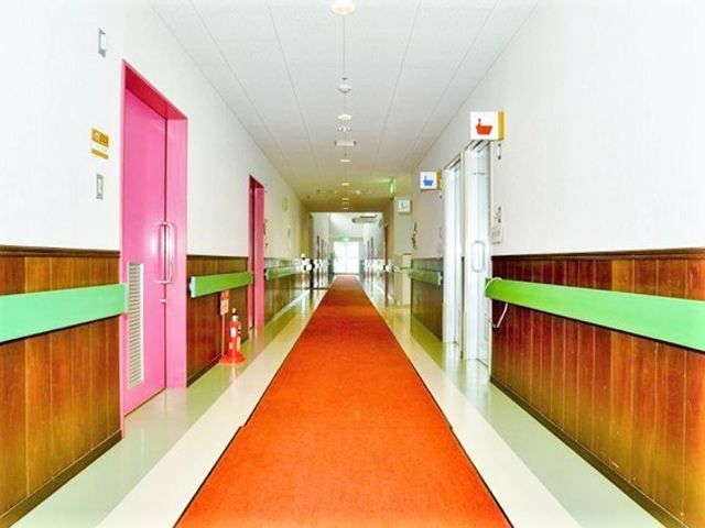 清潔な廊下の風景 廊下（２） 床は滑りにくい素材で、壁には手すりが設けられ、安心して歩行できる環境が整っています。カラフルな扉が明るい印象を与え、居心地の良さを演出しています。