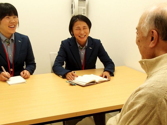 面談中の様子の居室 居室   柔らかな自然光が入る部屋で、スタッフが笑顔で相談に乗っています。親しみやすい雰囲気です。