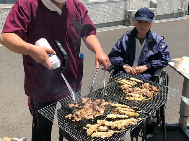 屋外でバーベキューをする様子 ケア スタッフが中庭でバーベキューをしており、車椅子の方も一緒に楽しんでいます。日差しの中、楽しいひと時を過ごせる場所です。