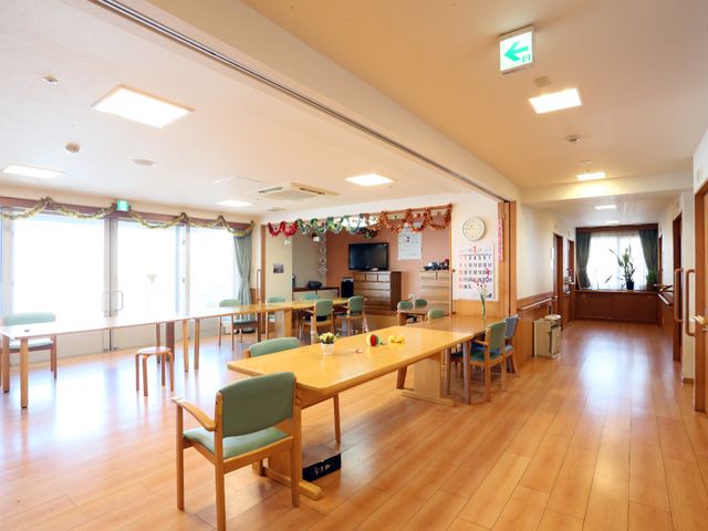 明るい共有ラウンジ空間 ダイニング ゆったりとした椅子配置のラウンジで、入居者が自由に過ごす姿が想像されます。正面の大きな窓から自然光がたっぷりと差し込んでいます。