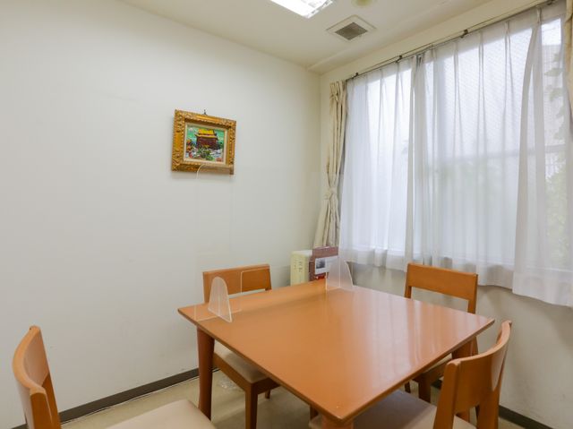 シンプルな居室のテーブル 相談室 ゆったりとした居室で、家庭的な雰囲気の食卓が設置されております。日の光が差し込むカーテン越しに、温かなひとときを提供しています。