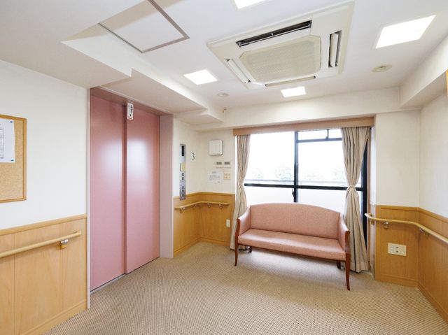 安らぎの居室エリア エレベーター前 手すり付きの廊下と落ち着いた色合いの応接セットが配された居室です。窓からの自然光で温もりのある空間をご提供しています。