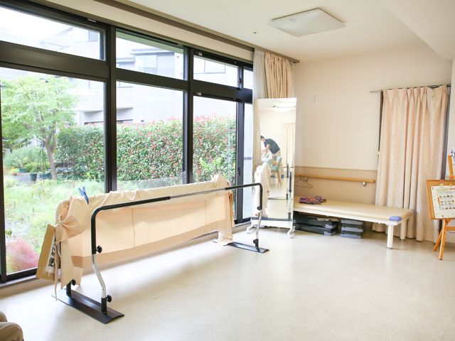 明るいガーデンビュー機能訓練室 機能訓練室 ゆとりのある機能訓練室では、窓辺で訓練の手助けをしているスタッフの姿が見え、穏やかな庭の景色も楽しめます。お仕事を通じて、利用者の方の心身のサポートができる環境です。