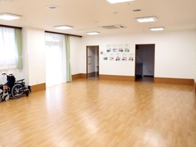 広々とした明るい多目的室 地域交流スペース ゆったりとした空間の多目的室は、壁には掲示物があり、様々な活動が行われる場としての可能性を秘めています。車椅子が置かれている様子からも、バリアフリーへの配慮がうかがえます。