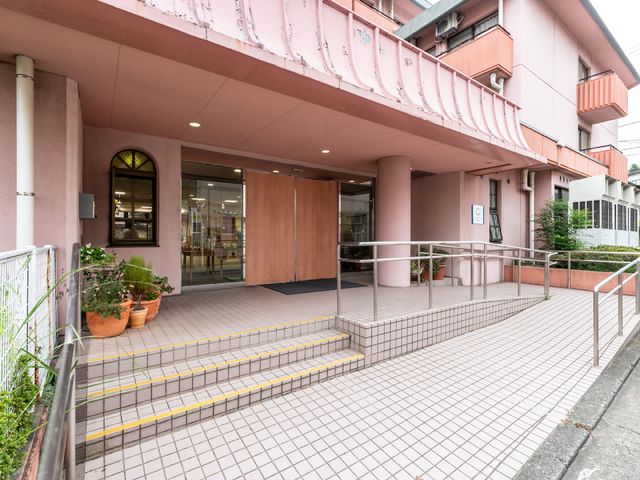 ピンクの建物エントランス 外観 手すり完備の広々としたエントランスです。明るさと開放感があるため、訪れる人々を温かく迎え入れます。