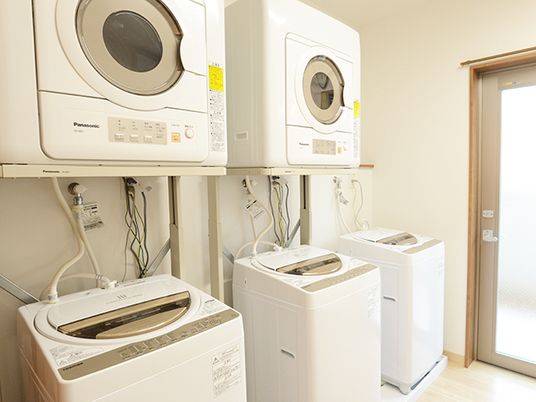縦型の洗濯機が３台が並び、上部にはドラム式の乾燥機が２台設置され、奥にはガラスのドアが付いている。きれいに掃除がされ清潔な空間。