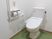 サムネイル 施設の写真 トイレは、白い壁にモスグリーンのシート張りの床になっている。洗浄機能付きの便器が据えられ、左の壁面には手すりが付けられている。