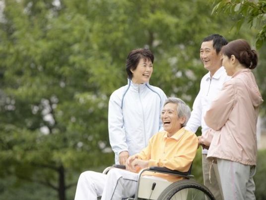 男性の入居者様が車椅子に乗っており、付き添っている３人の職員とともに、にこやかな表情を浮かべている。天気がいい日は散歩や外出を楽しめる。