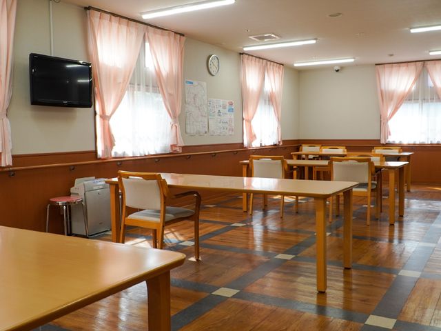 共有スペースの明るい一角 共有スペース ゆったりとした食堂では、木目の温かみを感じさせるテーブルが並び、明るく穏やかな時間を過ごせる空間が広がっています。壁掛けのテレビや掲示板も備え、情報共有や娯楽のための設備が整っています。