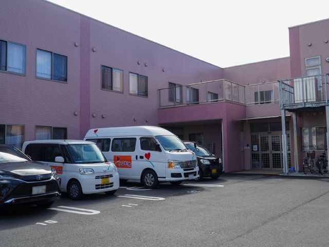 ピンクの壁の建物正面 外観 ピンク色の外壁が印象的な建物です。送迎車が配備されており、アクセスのしやすさが伺えます。