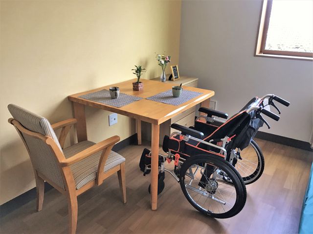 居室のテーブルと車椅子 ゆったりとした居室で、テーブルが置かれており日当たりも良好です。自己完結型の車椅子も完備され、快適な生活スペースが提供されています。