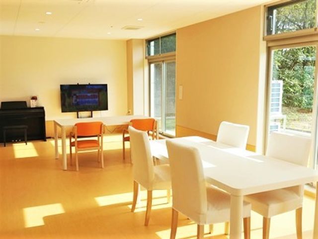リビングの明るい空間 リビング 採光豊かなリビングで、テレビやピアノが置かれ、憩いのひと時を提供しています。白いテーブルとオレンジの椅子が心地よい昼下がりを演出しています。