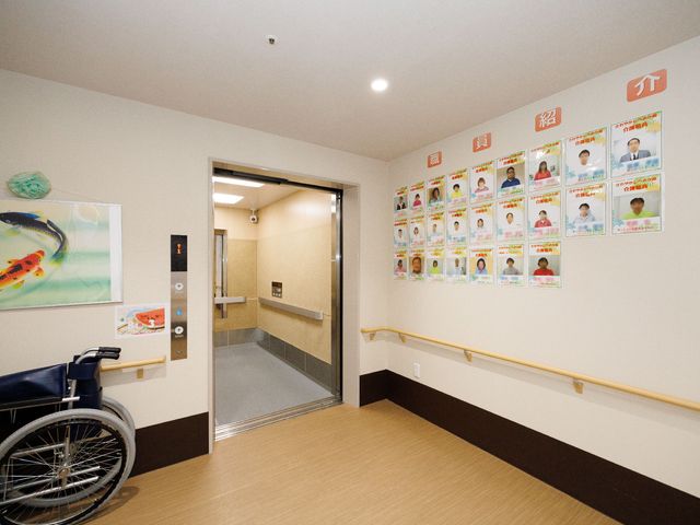 廊下に手すりと掲示物 エレベーター 清潔感のある廊下には、手すりが設けられ安全に配慮されています。壁にはスタッフの写真が掲示され、親しみやすい雰囲気を醸し出しています。