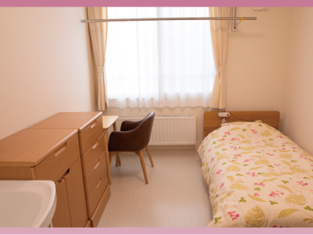 明るい居室のベッド 居室   窓からやわらかな光が入る、落ち着いた雰囲気の居室です。収納とデスクがあり、生活しやすい配置になっています。