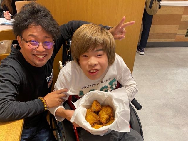 車椅子の若者と男性 和やかな雰囲気でスタッフが笑顔を交わし、リラックスした時間を過ごしている様子です。温かいコミュニケーションが感じられる職場環境をお求めなら、ぴったりかもしれません。