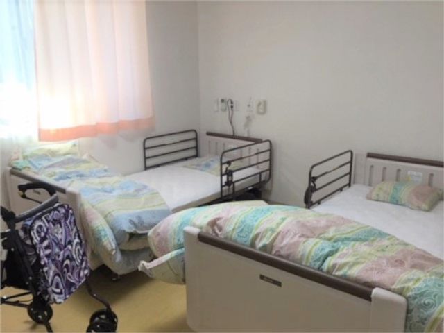 明るい個室の二つベッド 居室 明るく清潔感があるベッドのある部屋です。手前には車いすが置かれ、利用者の快適な生活空間が整っています。